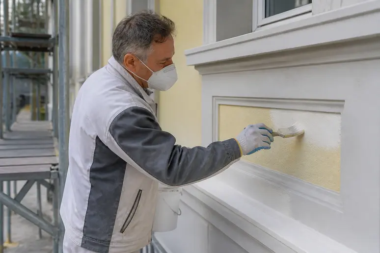 Maler renoviert Fassade eines Mehrfamilienhauses in Solingen mit Pinsel und Schutzausrüstung, Tageslichtaufnahmen.