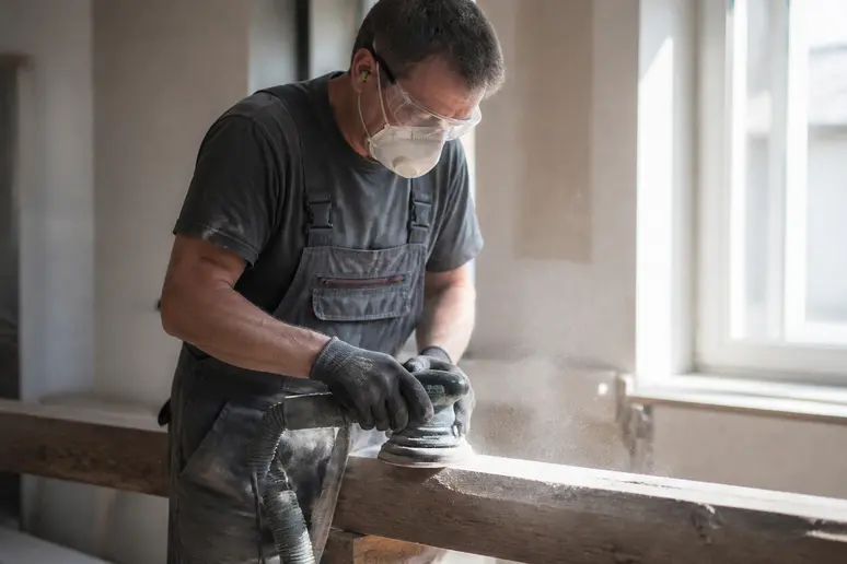 Ein Handwerker in Solingen schleift vorsichtig Holz mit Atemschutzmaske, Staubpartikel sind sichtbar.