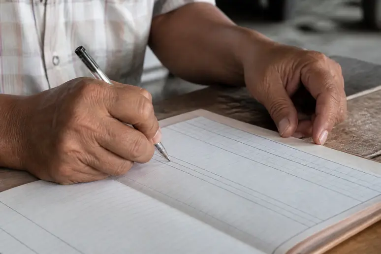 Hände füllen handschriftlich ein Kassenbuch mit Datum, Beschreibung, Einnahmen und Ausgaben.